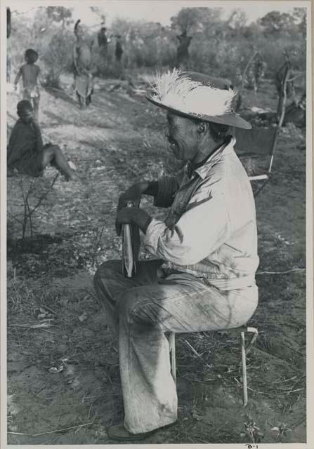 Wilhelm Camm sitting in a chair in profile, people in the background
