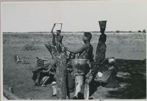 Group of women at water trough, two women standing, carrying buckets on their heads

