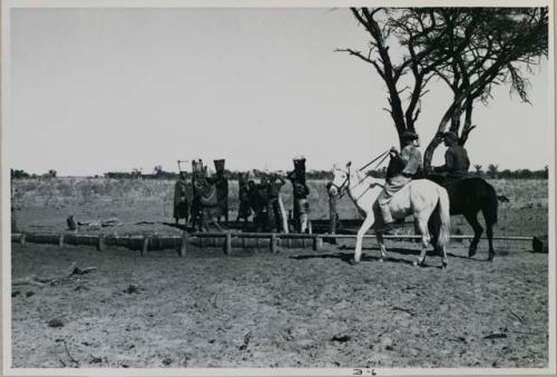 Two men on horses; a group of women by the water trough carrying buckets on top of their heads
