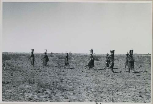 Group of women walking in a line, carrying buckets on top of their heads
