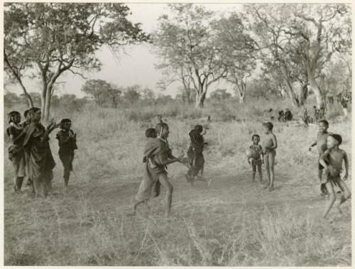 "Women's Ball Game": Girls playing N!owa t/ama (melon tossing game) / tamah n!o’an (ball game); four girls to the left are singing and clapping and an unidentified, visiting girl in the center carries a child on her back while she plays; other children present in the background (print is a cropped image)