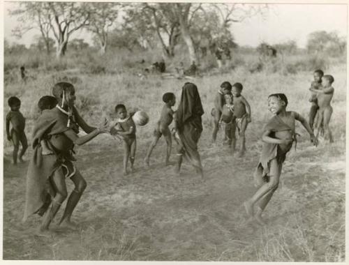 "Women's Ball Game": Girls playing N!owa t/ama (melon tossing game) / tamah n!o’an (ball game); an unidentified, visiting girl carries a child on her back as she plays; other children present in the background (print is a cropped image)