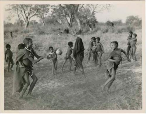 "Women's Ball Game": Girls playing N!owa t/ama (melon tossing game) / tamah n!o’an (ball game); an unidentified, visiting girl carries a child on her back as she plays; other children present in the background (print is a cropped image)