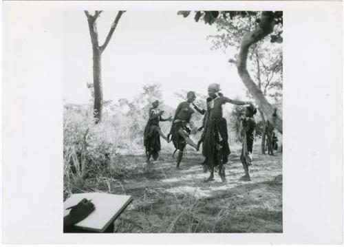 "The 1957-8 Games": Group of women and girls playing Tcxai Djxani (a dancing game), dancing in a line, with N!ai at the end of the line, clapping (print is a cropped image)