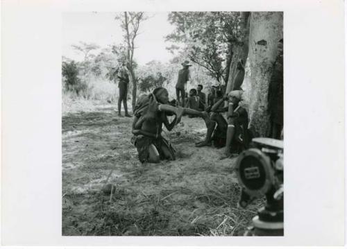 "The 1957-8 Games": Group of women and girls playing Tcxai Djxani (a dancing game) at a tree, with one girl putting another girl on her shoulders, John Marshall filming them (print is a cropped image)