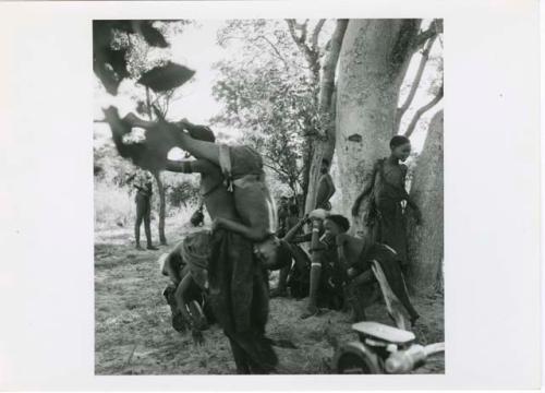 "The 1957-8 Games": Group of women and girls playing Tcxai Djxani (a dancing game), with a girl carrying a child over her back, John Marshall filming them (print is a cropped image)