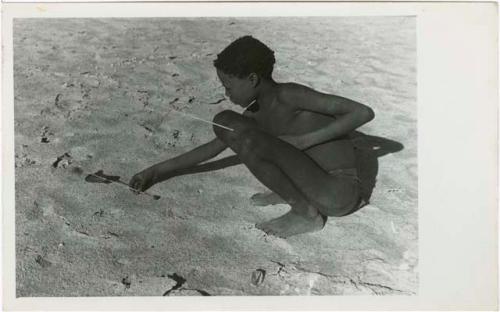 "Djani": Ju/'hoan boy squatting with djani bird (helicopter toy) (print is a cropped image)