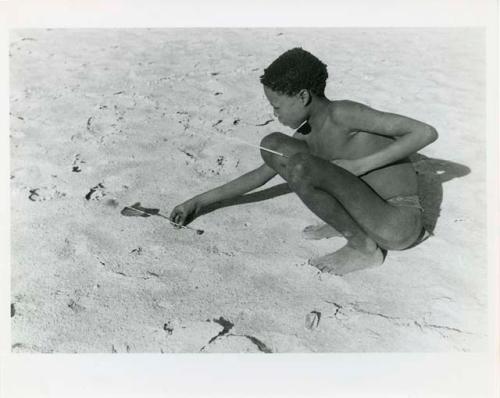 "Djani": Ju/'hoan boy squatting with djani bird (helicopter toy) (print is a cropped image)