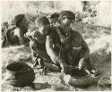 "The 1955 study of groups with children": Di!ai with a bowl, a knife, and a pot of meat beside her, with Debe leaning over her shoulder watching (print is a cropped image)