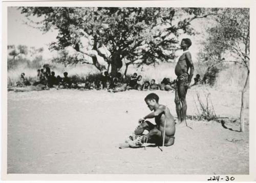 "Children": /Ti!kay sitting and putting on his dance rattles, Gau standing beside him, with people sitting under a tree in the background (print is a cropped image)