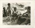 "Photos of Children": Unidentified person and Bau, who is playing with a digging stick to the left, Debe watching /Gaishay cut a piece of meat with a knife, and /Naoka looking through "Gao Medicine's" hair for lice; part of Elizabeth Marshall Thomas's child study (print is a cropped image)