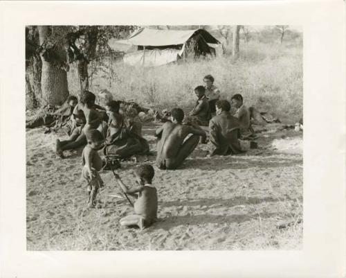 "Photos of Children": Debe and /Gaishay sitting near a group of people, including ≠Toma (wearing a coat), with !Ungka Norna playing near the group, Elizabeth Marshall Thomas observing (print is a cropped image)