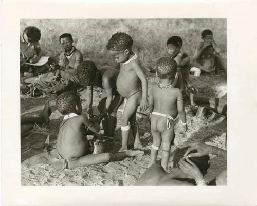 "Photos of Children": !Ungka Norna watching /Gaishay and Bau sharing food, Elizabeth Marshall Thomas taking notes, and other unidentified people; part of Elizabeth Marshall Thomas's child study (print is a cropped image)