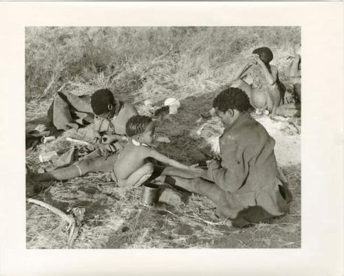 "Photos of Children": !Ungka Norna seated between !U and ≠Toma who holds a piece of meat and a knife with two unidentified people in the background (print is a cropped image)