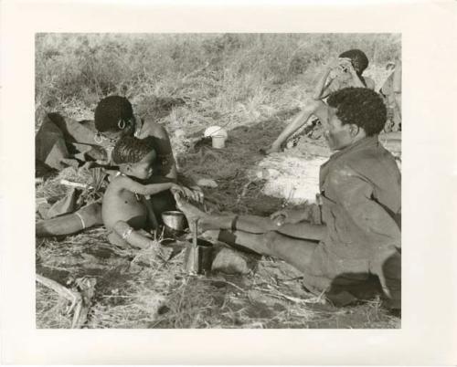 "Photos of Children": !Ungka Norna holding a piece of meat, seated between !U and ≠Toma with two unidentified people in the background (print is a cropped image)