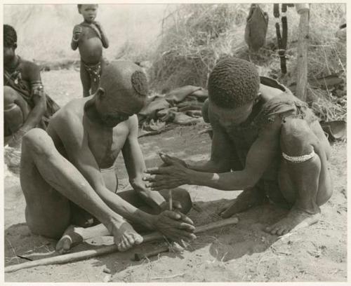 Dam and ≠Gao creating a fire with firesticks; unidentified people in the background (print is a cropped image)