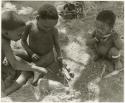 Unidentified boy to the left, "Little ≠Gao" playing with a handmade toy automobile made from veldkos, and Debe ("Gao Medicine" and Di!ai's son) eating something (print is a cropped image)
