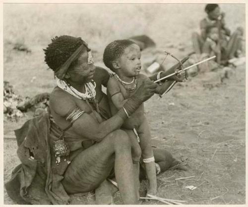 Di!ai showing her son, Debe, how to hold a toy bow and arrow (print is a cropped image)