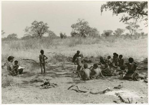 Women and children sitting in the shade while Elizabeth Marshall Thomas holds a child, distant view (print is a cropped image)