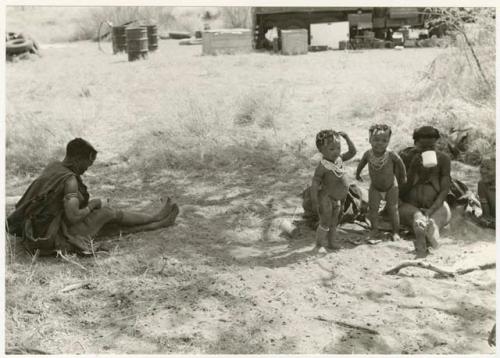 Khuan//a sitting to the left, her youngest son ≠Toma standing with an unidentified girl, and two unidentified women with the expedition camp in the background (print is a cropped image)