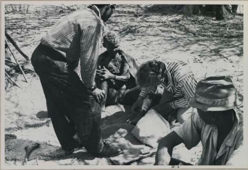 Ngani and Lorna Marshall bending over Xama, treating her leg; person sitting and watching them

