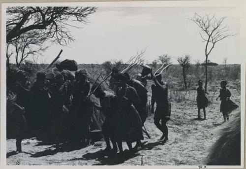 Group of people performing the Eland Dance; group of women clapping; men holding wooden horns to their heads



