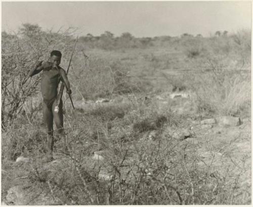 Distant view of "Crooked /Qui" standing in brush, with his quiver, spear, bow, and arrow (print is a cropped image)