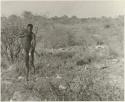 "Crooked /Qui" standing in brush, with his quiver, spear, bow, and arrow, distant view (print is a cropped image)