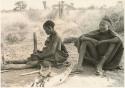 "Old /Gasa" sitting, legs straight out, pounding something with mortar and pestle; her grandson sitting beside her (print is a cropped image)