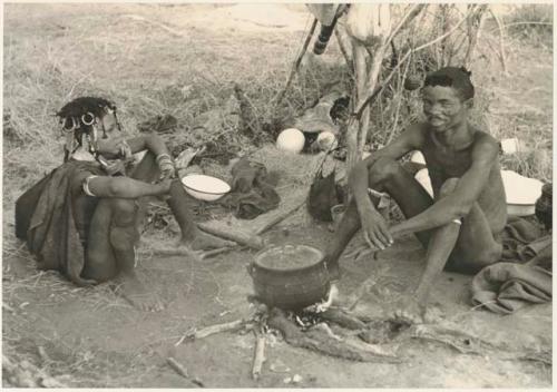 "Gao Music" and his wife, ≠Nisa, sitting at their living place with the iron pot on the fire (print is a cropped image)