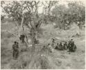 Group of Ju/'hoansi sitting in a cluster under a tree, with a woman holding a baby standing apart (print is a cropped image)