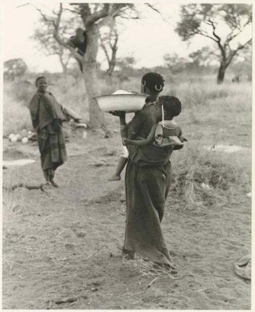 !U carrying a large basin of mealie meal, with a child tied to her back, seen from behind (print is a cropped image)