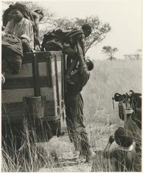 Kernel Ledimo lifting !U down from a truck, with other Ju/'hoansi in the background (print is a cropped image)