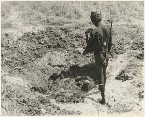 "/Gao Music" standing by a waterhole, seen from behind (print is a cropped image)