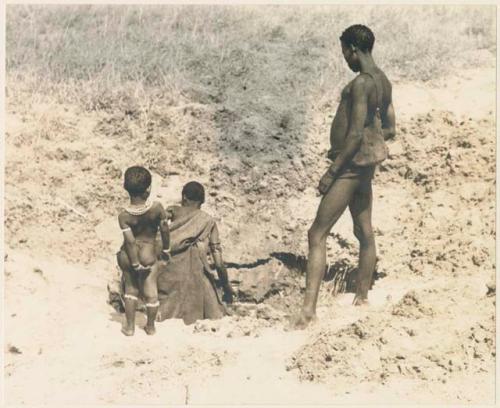 Woman filling eggshell in a waterhole; "Crooked /Qui" and an unidentified child standing behind her (print is a cropped image)