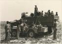 Expedition members filling Jeep gasoline tank from barrel in GMC trucks, accompanied by a group of Ju/'hoansi (print is a cropped image)