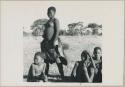 Tsetse girl who is a Makoba of Okavango group, standing next to a group of other people sitting (print is a cropped image)