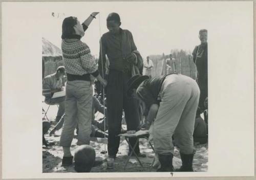 Man having his height measured by Deborah Marshall; Lorna Marshall bending over and taking notes (print is a cropped image)