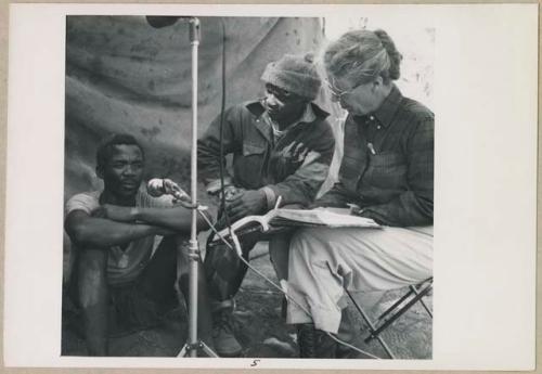 Man speaking a list of words into a microphone; Kernel Ledimo and Lorna Marshall sitting next to him (print is a cropped image)