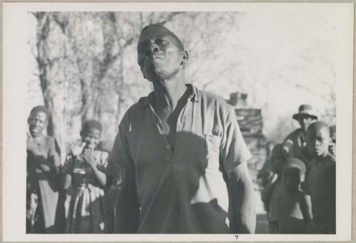 Man dancing, close-up; group of people singing and clapping in the background (print is a cropped image)