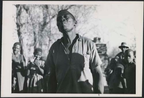 Man dancing, close-up; group of people singing and clapping in the background (print is a cropped image)