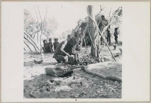 Kumsa cutting meat, with women and children in background (print is a cropped image)