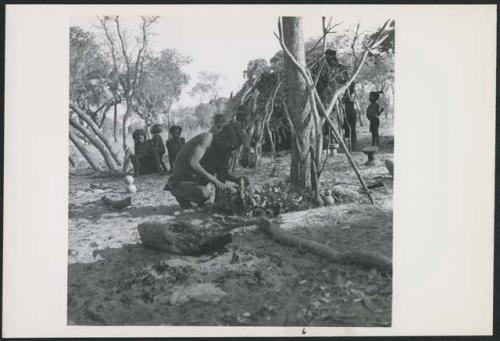 Kumsa cutting meat, with women and children in background (print is a cropped image)