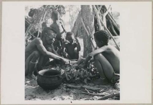 Kumsa cutting something next to a tree, children watching (print is a cropped image)