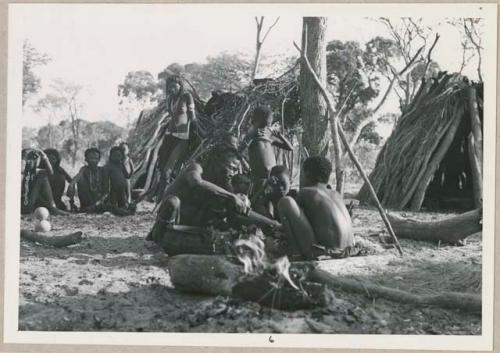 Kumsa cutting something next to a tree, children watching; other women and children in the background (print is a cropped image)