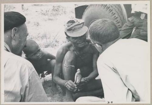 "/Gao Overalls" holding a bird with O.P.M. Prozesky and Nicholas England sitting across from him, seen from behind (print is a cropped image)