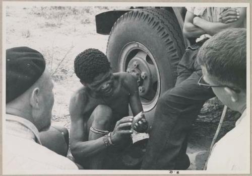 "/Gao Overalls" holding a bird sitting with O.P.M. Prozesky and Nicholas England sitting across from him, seen from behind (print is a cropped image)