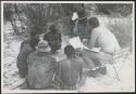 Wilhelm Camm and Nicholas England in chairs with a sound recorder interviewing five men sitting on the ground (print is a cropped image)