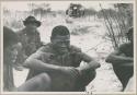 Four men sitting on the ground; truck in the background (print is a cropped image)