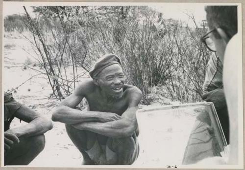 Wilhelm Camm and Nicholas England in chairs with a sound recorder interviewing two men sitting on the ground, close-up (print is a cropped image)